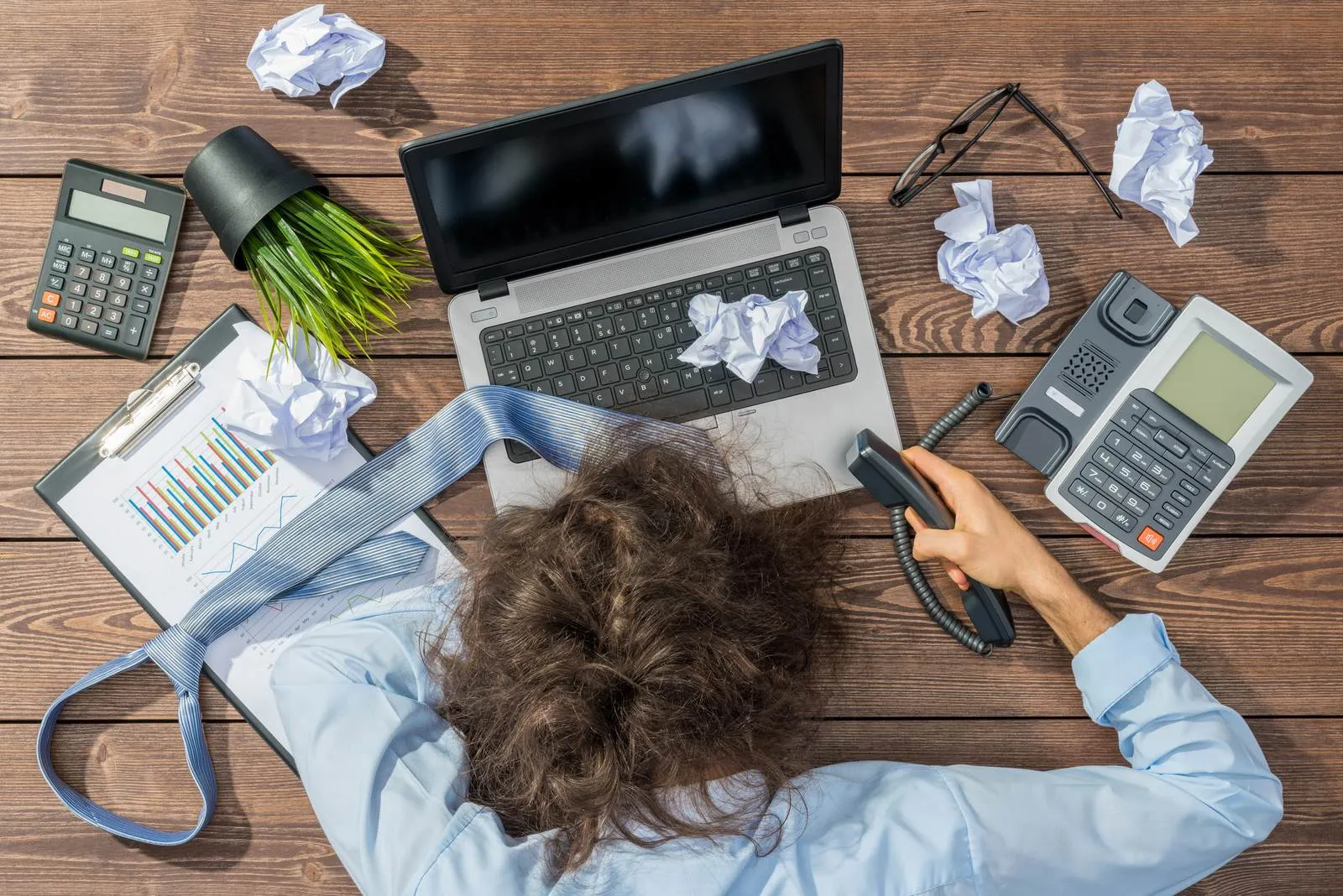 A property manager slumped over a desk cluttered with a laptop, a phone, printed spreadsheets, a calculator and crumpled notes