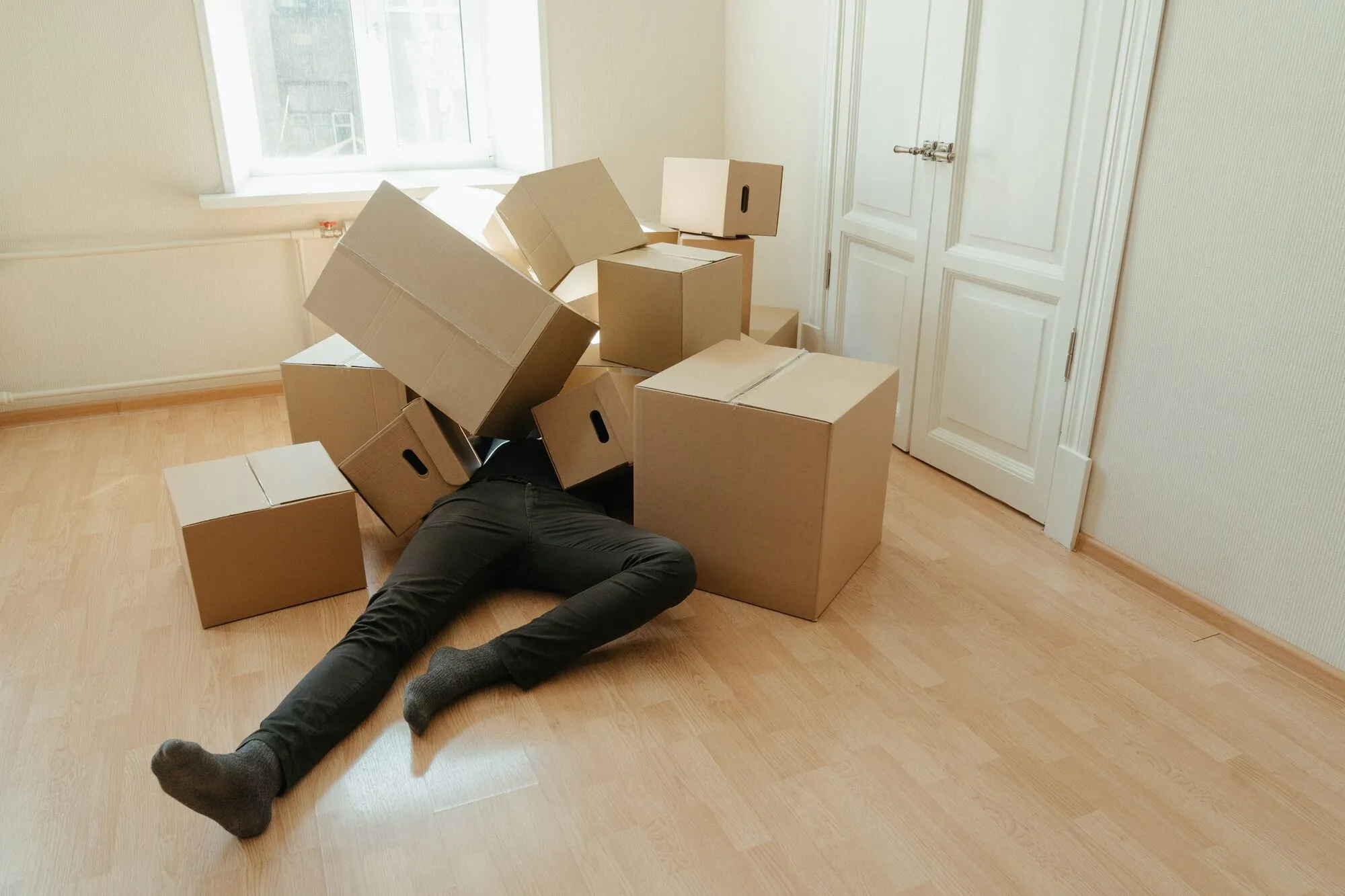 A person lying on the floor of an empty apartment, buried under a pile of cardboard moving boxes, representing a first-time landlord overwhelmed by avoidable mistakes
