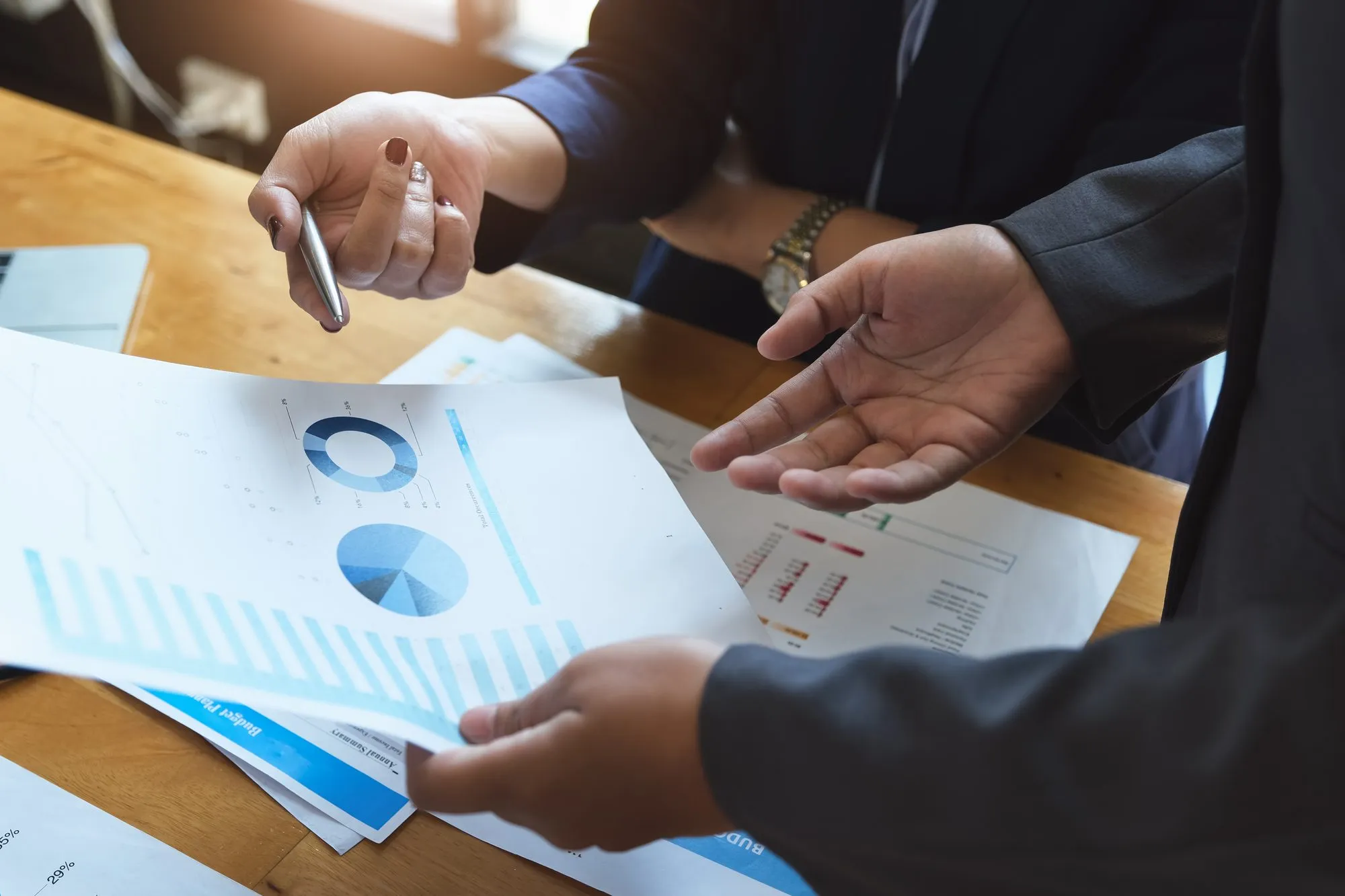 Two people in business attire going over financial documents at a desk, one pointing at the numbers with a pen and the other gesturing with open palms