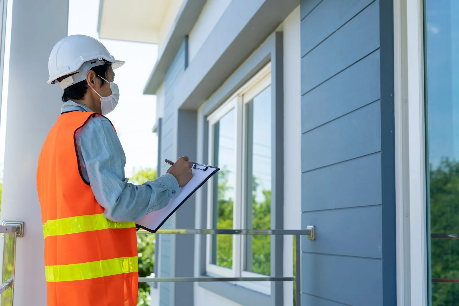 An inspector in a white hard hat and orange safety vest making notes on a clipboard while checking the exterior windows of a rental property